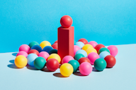 A vibrant composition of colourful balls surrounding a red pillar topped with a red ball, set against a bright blue background.
